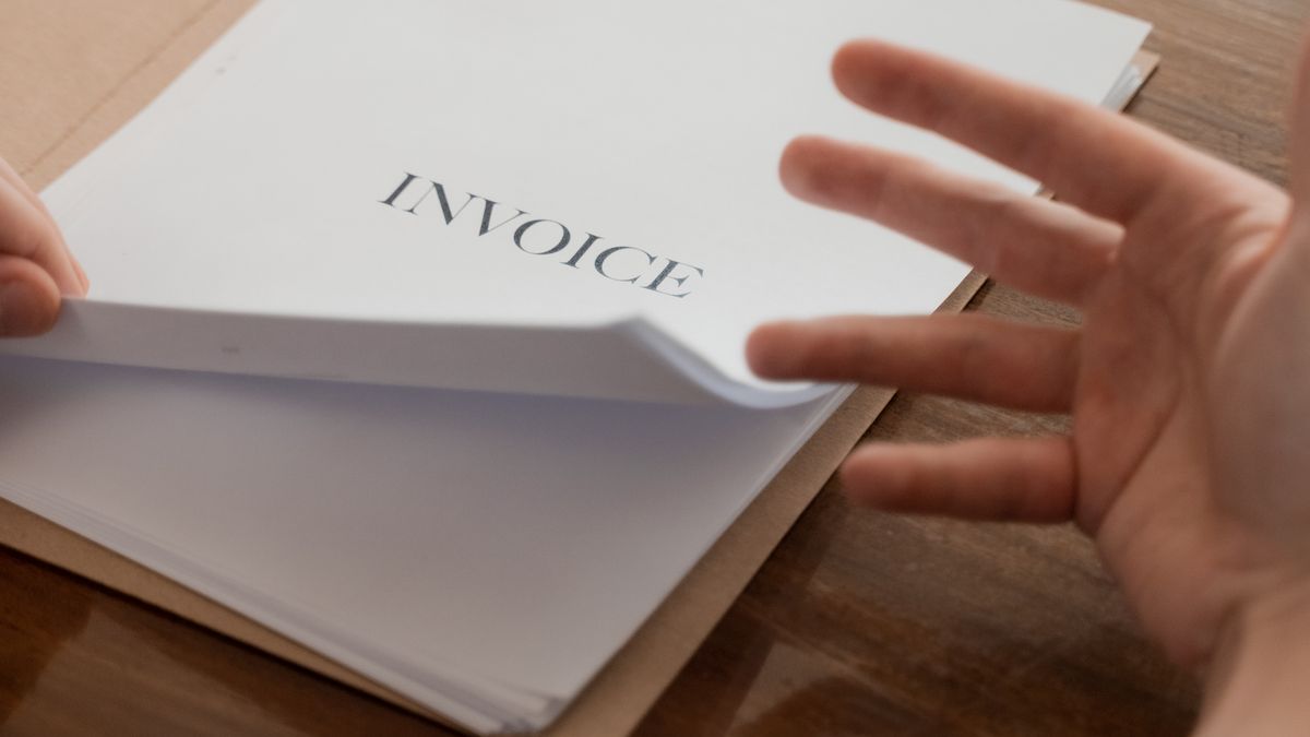Manos pasando las hojas de un taco de facturas impresas con la palabra INVOICE en la portada, sobre una mesa de madera