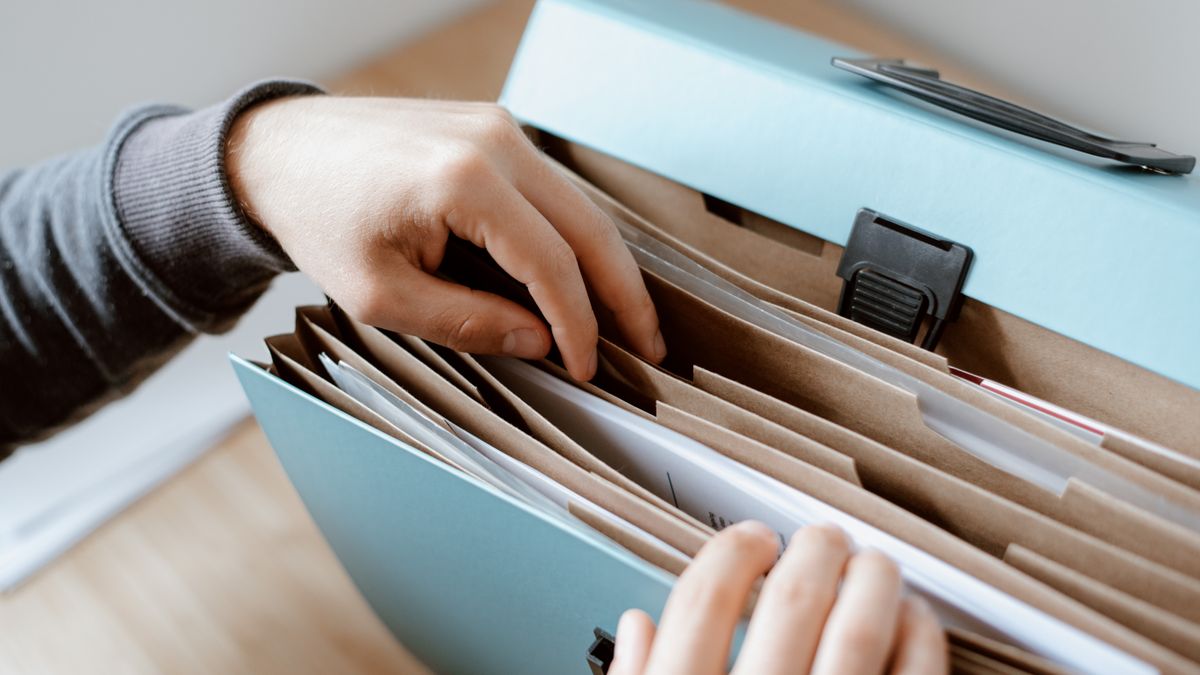 Manos abriendo una carpeta de fuelle azul llena de documentos separados por pestañas de cartón, sobre una mesa de madera clara