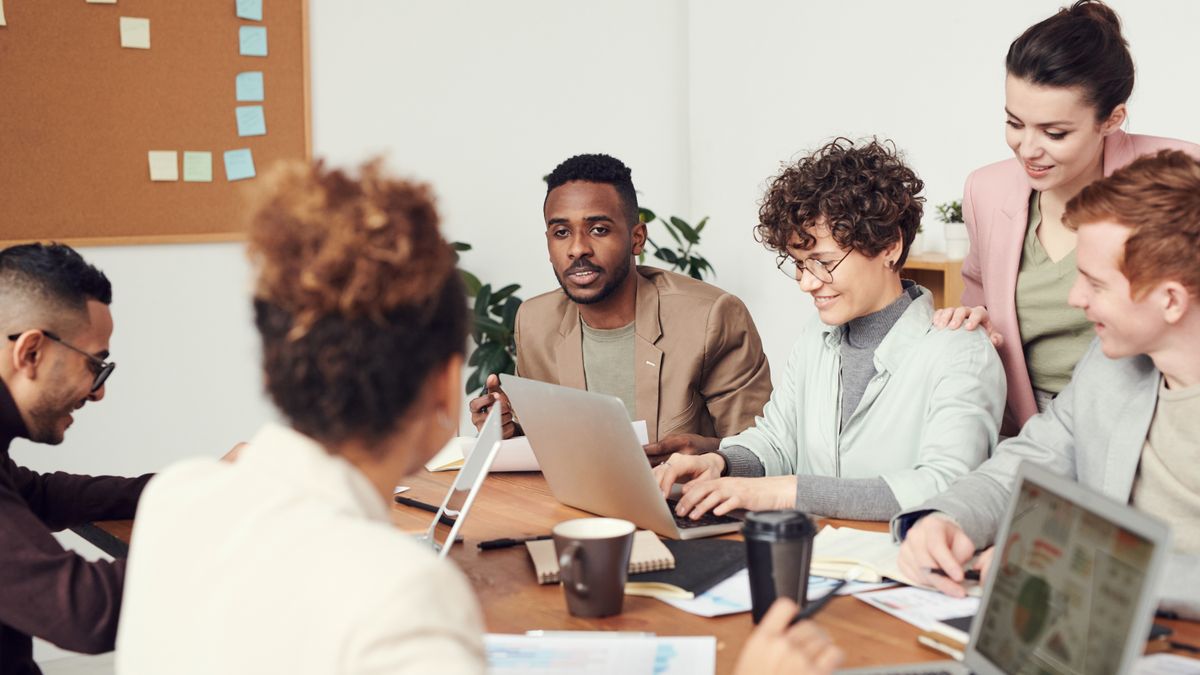 Grupo de compañeros de trabajo reunidos alrededor de una mesa mirando la pantalla de un portátil, con un tablón de notas adhesivas al fondo
