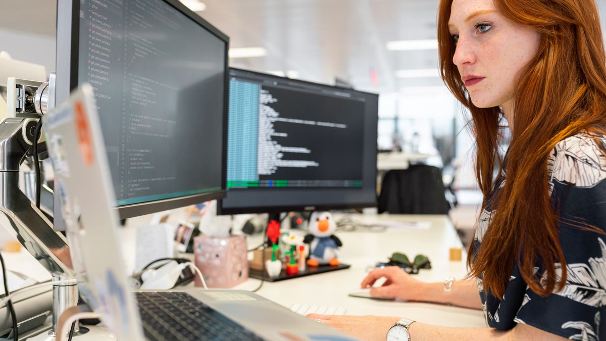 Persona trabajando en una oficina abierta frente a dos monitores con código y una consola en pantalla