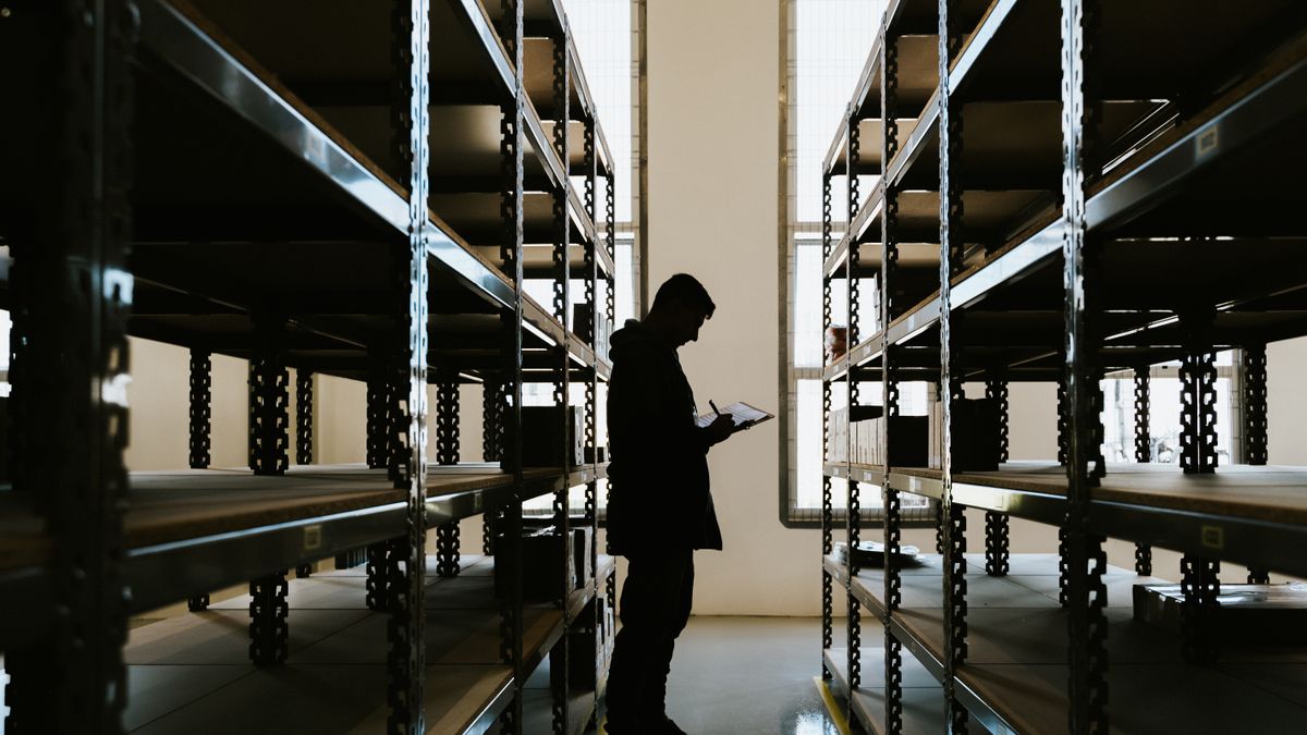 Person holding a clipboard and writing between two rows of metal warehouse shelving, backlit