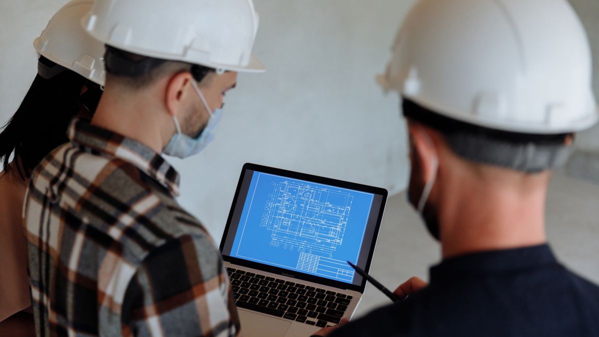 Three people in white hard hats and face masks look at a laptop showing a blue floor plan, inside a building site