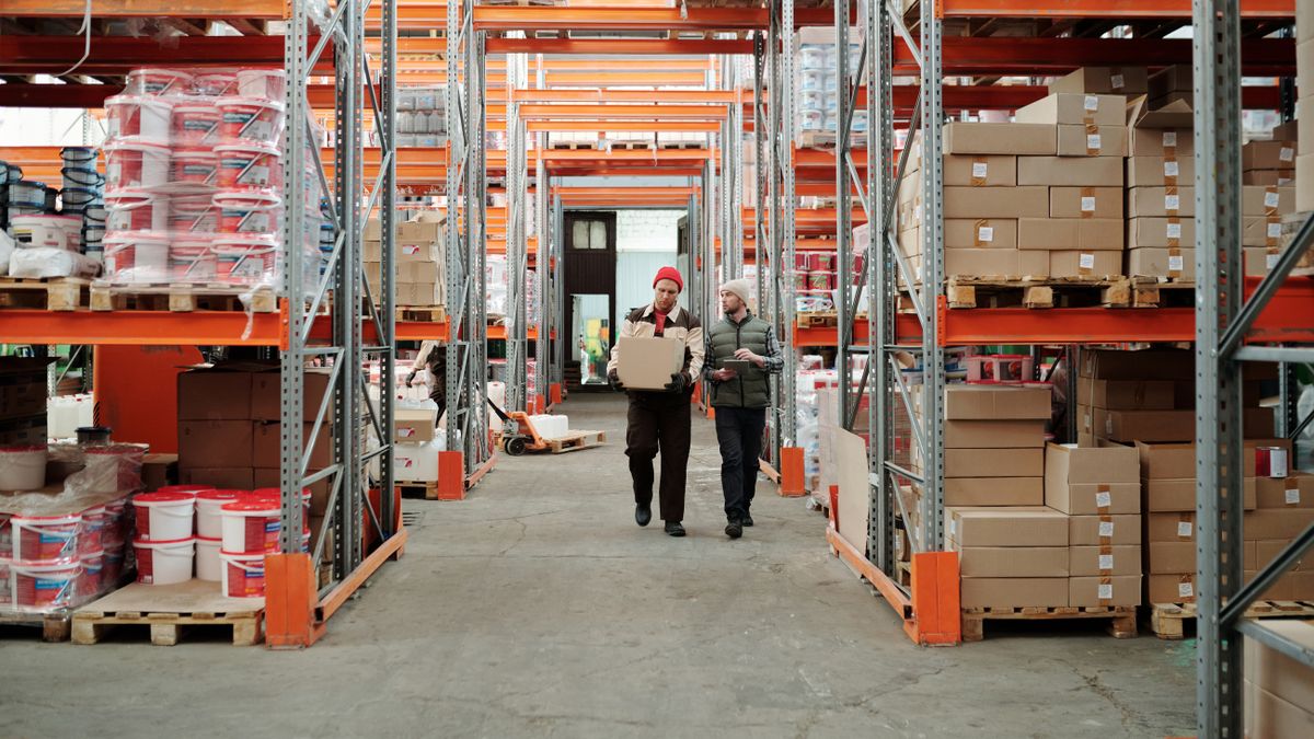 Two workers walk down the central aisle of a warehouse with orange racking full of boxes and pallets; one carries a box, the other a tablet