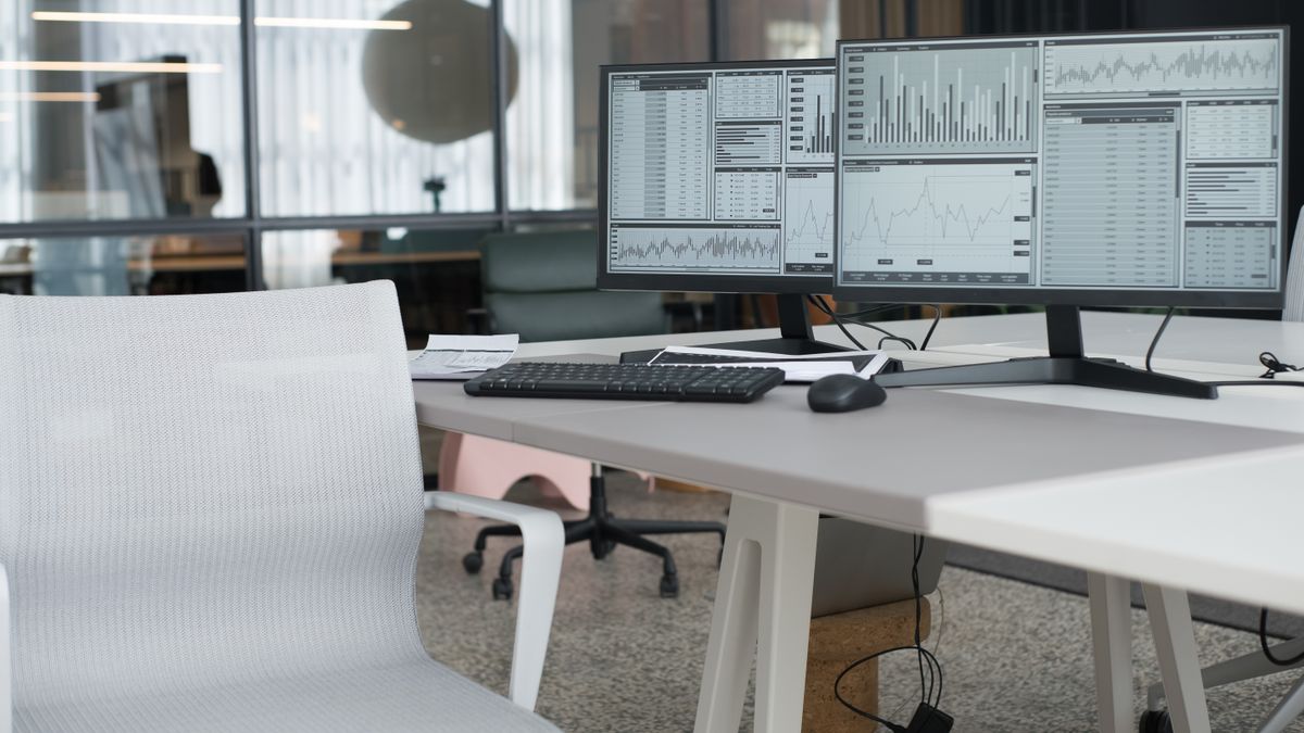 Empty office desk with two monitors switched on showing data tables and charts, a keyboard and a mouse on the surface