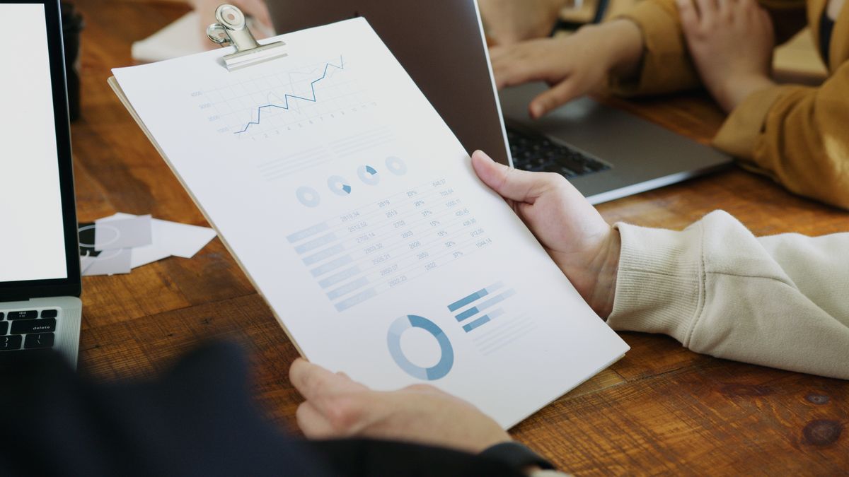 Manos sosteniendo un portapapeles con un informe impreso de gráficos de líneas y quesos junto a un portátil sobre una mesa de madera