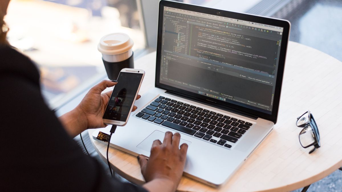Persona sentada ante un portátil con un editor de código abierto en pantalla, sujetando un móvil, con un café y unas gafas sobre la mesa