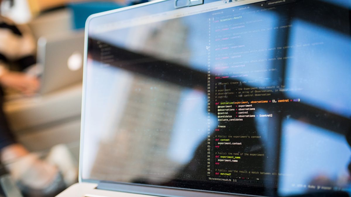 Laptop screen showing colour-highlighted source code in an editor, with an office window reflected in the glass