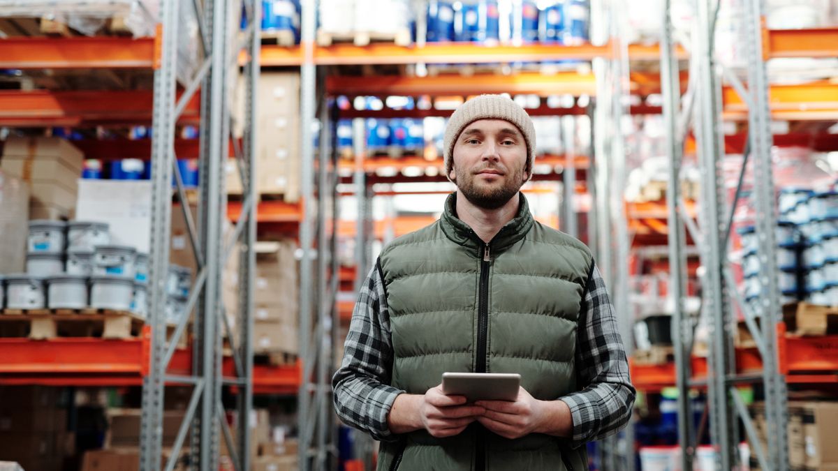 Warehouse worker in a beanie and gilet holding a tablet in an aisle lined with shelves full of boxes and tubs