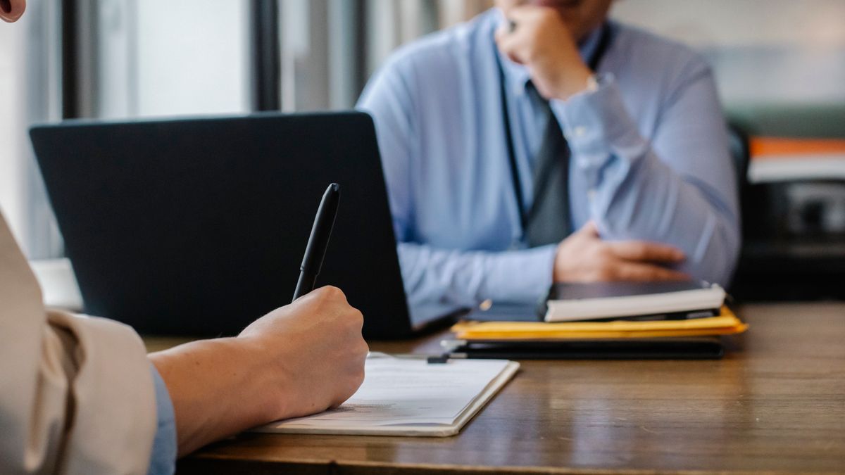 Two people at a meeting table; one takes handwritten notes in a notebook next to a laptop while the other listens