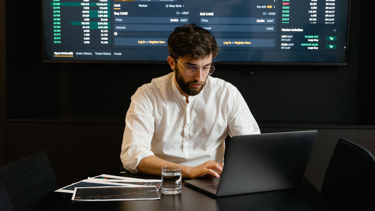Man working on a laptop in a dark room, with a large screen behind him full of figures and price quotes in green and red