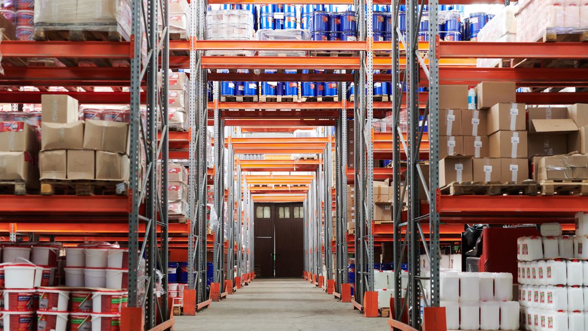 Pasillo de un almacén con estanterías metálicas llenas de cajas, palés y envases apilados hasta arriba
