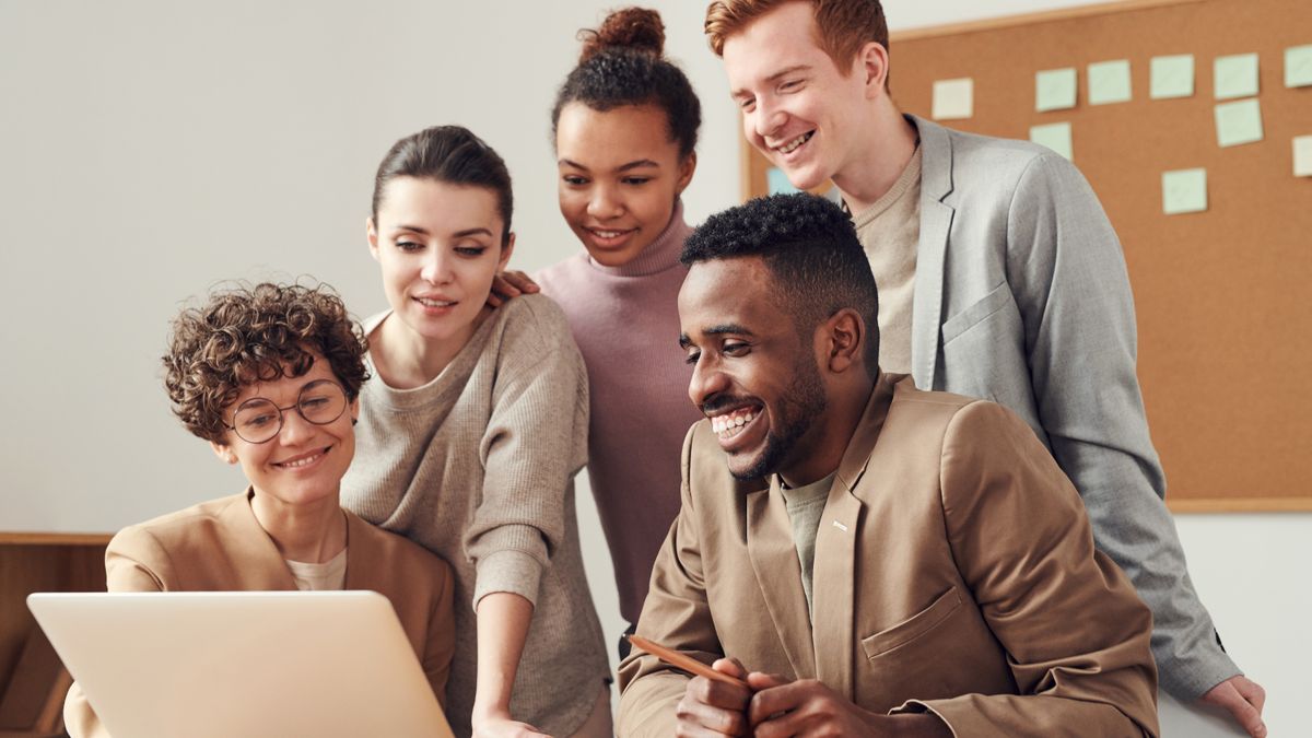Five smiling colleagues looking together at a laptop screen in an office